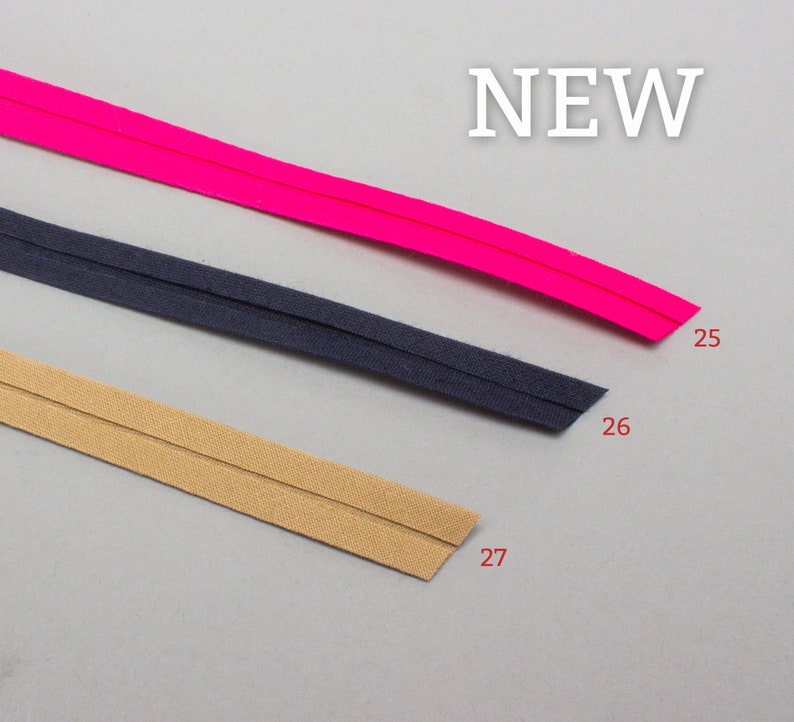 May include: Three different colors of bias tape, pink, navy blue, and beige. The tape is folded in half and has a seam down the center. The tape is labeled with numbers 25, 26, and 27.
