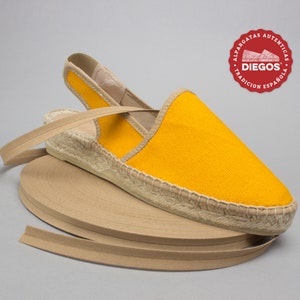 May include: Yellow espadrille sandals with a brown jute sole and a brown ribbon tie. The sandals are on a roll of brown ribbon. The brand name "DIEGOS" is on a red circle with the text "Alpargatas Aut&eacute;nticas Tradici&oacute;n Espa&ntilde;ola".