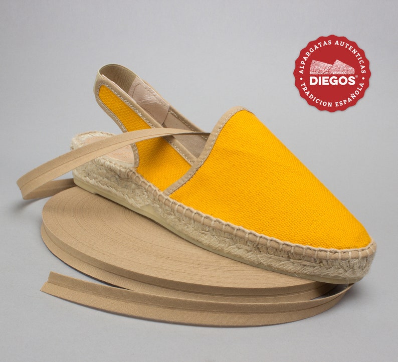 May include: Yellow espadrille sandals with a brown jute sole and a brown ribbon tie. The sandals are on a roll of brown ribbon. The brand name "DIEGOS" is on a red circle with the text "Alpargatas Aut&eacute;nticas Tradici&oacute;n Espa&ntilde;ola".