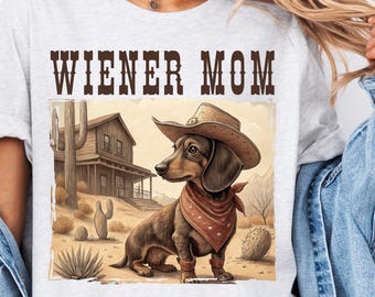 May include: A white t-shirt with the text "WIENER MOM" printed in brown letters. The shirt features a cartoon image of a dachshund wearing a cowboy hat and bandana in a desert setting.