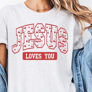 Op de afbeelding: Wit T-shirt met een rode grafische afbeelding waarop "JESUS LOVES YOU" staat in een varsity-lettertype. De letters zijn gevuld met een patroon van rode kersen en harten.