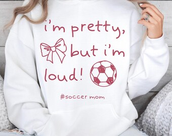 May include: White hoodie with a drawstring and a front pocket. The hoodie features the text "I'm pretty, but I'm loud!" in burgundy, along with a bow and a soccer ball graphic. The hashtag "#soccer mom" is also included.