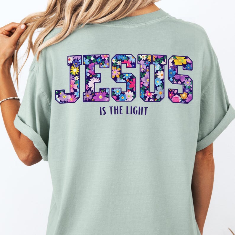 May include: A light green t-shirt with the word "JESUS" in large, floral-filled letters across the back. Below the word "JESUS" is the phrase "IS THE LIGHT" in white text. The shirt has rolled-up sleeves.