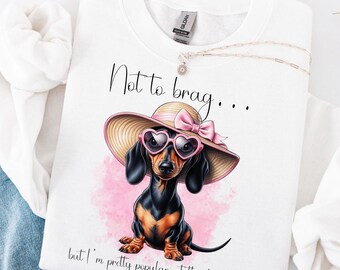 May include: White t-shirt with a black and brown dachshund wearing pink heart-shaped sunglasses and a pink bow on a straw hat. The text on the shirt reads "Not to brag... but I'm pretty popular at the dog park."