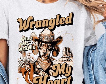 May include: A white t-shirt with a brown dachshund wearing a cowboy hat and bandana. The text "Wrangled Wild West Weenie My Heart" is printed on the shirt.