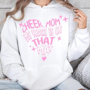 Può includere: Felpa con cappuccio bianca con un design a forma di cuore rosa che recita "Cheer Mom, My Heart Is On That Mat". Il design include stelle e un cuore. La felpa ha una tasca frontale e un cappuccio.