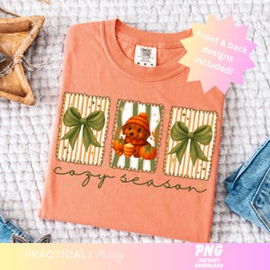 May include: Peach-colored t-shirt with a fall-themed design. The design includes a dog wearing a hat, pumpkins, and green bows. The text "cozy season" is written in a script font. The shirt also includes the text "Front & back designs included!"