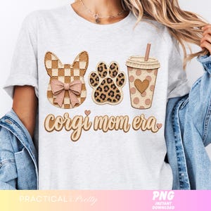May include: A light gray t-shirt featuring a corgi-themed design. The design includes a corgi head, a paw print, and a coffee cup, all in a neutral color palette. The text "corgi mom era" is written in a script font.