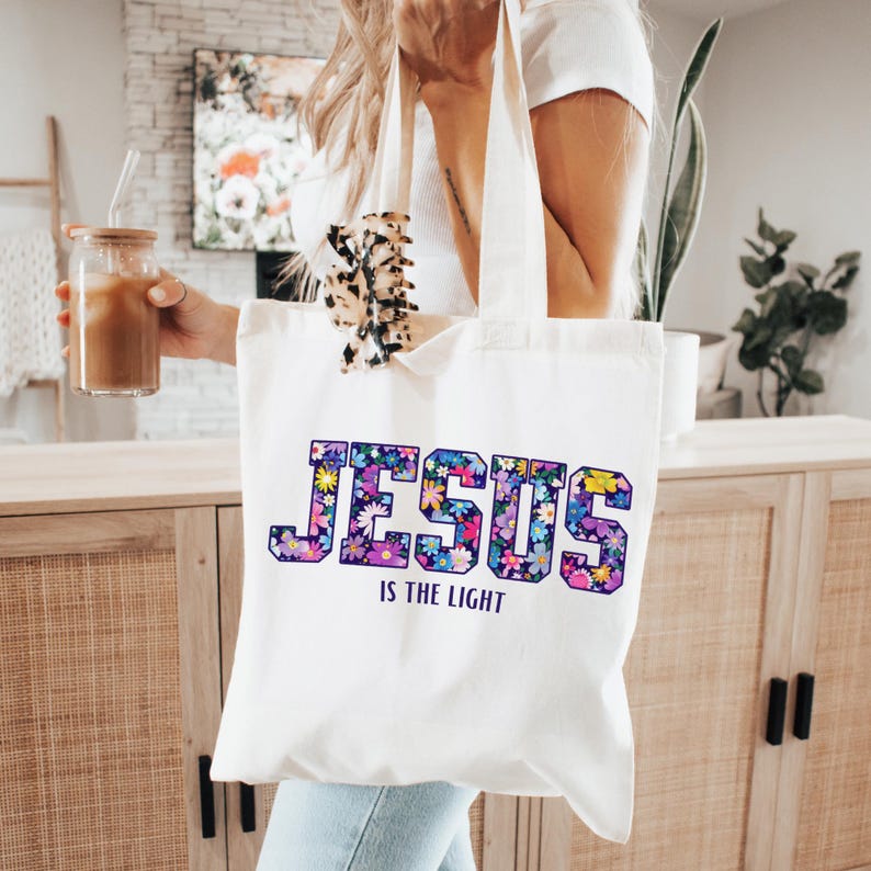 May include: A white tote bag with the words "JESUS IS THE LIGHT" in a floral design. The bag is held by a person. The tote bag is made of a light-colored fabric.