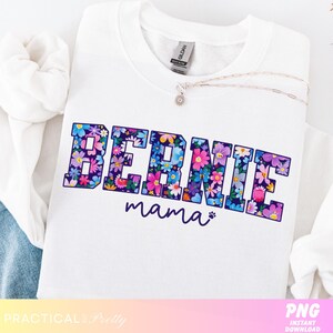 May include: White sweatshirt with the text "BERNIE mama" in a floral design. The letters are filled with colorful flowers in shades of blue, pink, and purple. A silver necklace with a small pendant is draped across the shirt.
