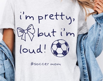 May include: A light gray t-shirt with the text "I'm pretty, but I'm loud!" in dark blue script. A bow and a soccer ball are drawn next to the text. The hashtag "#soccer mom" is below the text.