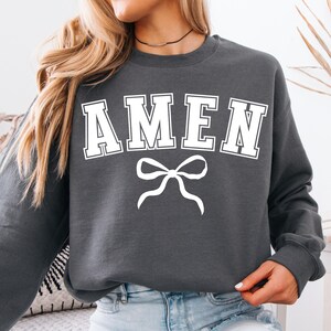 Op de afbeelding: Een donkergrijs sweatshirt met het woord "AMEN" in wit gedrukt, met een witte strik eronder.