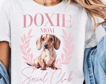 May include: Light gray t-shirt with a pink graphic. The graphic features the words "DOXIE MOM Social Club" and a detailed illustration of a dachshund dog. The words "LONG DOG LOVE CLUB" are at the bottom of the graphic.