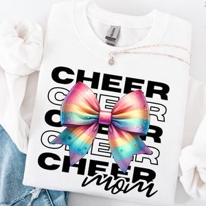 May include: White sweatshirt with the text "CHEER mom" in black. A large rainbow-colored bow is centered on the shirt.