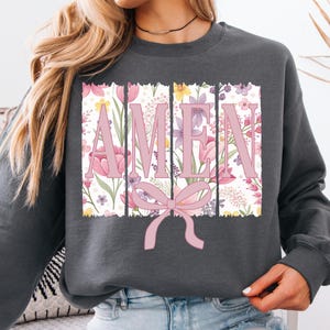 Op de afbeelding: Een antracietkleurig sweatshirt met een bloemenprint en het woord "AMEN" in roze letters. Een roze strik bevindt zich in het midden onder het woord.