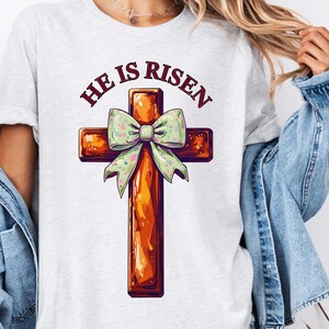 Op de afbeelding: Een wit T-shirt met een bruin houten kruis versierd met een groene strik. De tekst "He is Risen" is in rood boven het kruis gedrukt.