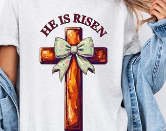 Peut inclure: Un t-shirt blanc avec une croix en bois brun ornée d'un nœud vert. Le texte "He is Risen" est imprimé en rouge au-dessus de la croix.