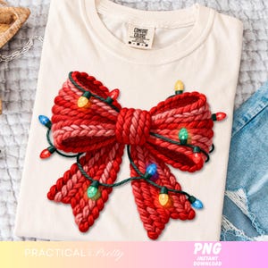 May include: Cream-colored t-shirt featuring a large, knitted red bow with a string of colorful Christmas lights. The bow is detailed with varying shades of red and pink. The shirt has a Comfort Colors tag.