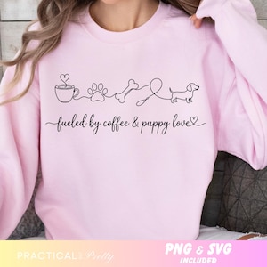 May include: A light pink sweatshirt with a black line drawing of a coffee cup, paw print, bone, and dachshund dog. The text "fueled by coffee & puppy love" is written below the drawing. The sweatshirt is a casual style.