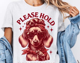 May include: A white t-shirt with a red graphic of a dachshund wearing sunglasses and a red bandana. The text "PLEASE HOLD SARCASM LOADING" is printed in red on the shirt.