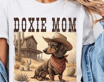 May include: A white t-shirt with the text "DOXIE MOM" printed in brown letters. The shirt features a cartoon image of a dachshund wearing a cowboy hat and bandana in a desert setting.