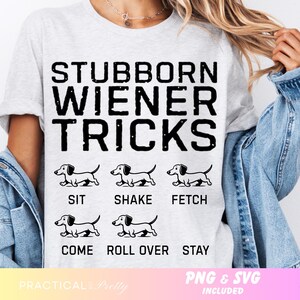 May include: A light grey t-shirt featuring the text "STUBBORN WIENER TRICKS" in black, bold font. Below the text are illustrations of dachshunds performing tricks. The shirt also includes the phrases "SIT", "SHAKE", "FETCH", "COME", "ROLL OVER", and "STAY".