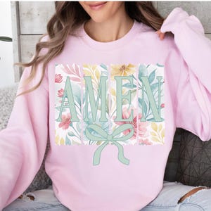 Puede incluir: Sudadera rosa con un diseño floral y la palabra "AMEN" en letras verdes con un lazo verde.