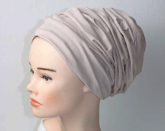 May include: A light beige turban-style head covering made of soft, stretchy fabric. The fabric is gathered and draped to create a loose, comfortable fit.