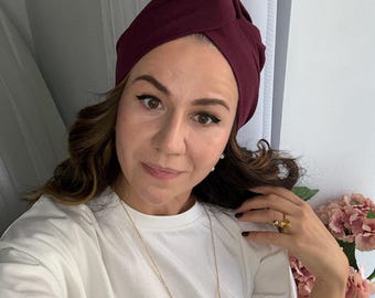May include: A burgundy turban-style head covering. The fabric is a solid color and appears to be soft. The turban is worn on the head. The person in the image is wearing a white shirt and a gold necklace.