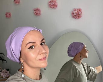 May include: A person wearing a light purple turban and a light green t-shirt. The person is standing in front of a mirror and looking at the camera. The mirror is reflecting the person's back.