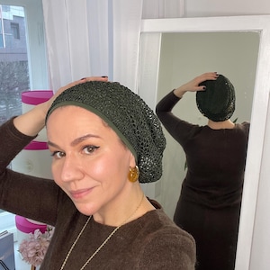 May include: A dark green crocheted beanie with a matching trim is worn. The person in the image is wearing a brown sweater and gold-coloured earrings. A gold necklace is also visible. The person is standing in front of a mirror.