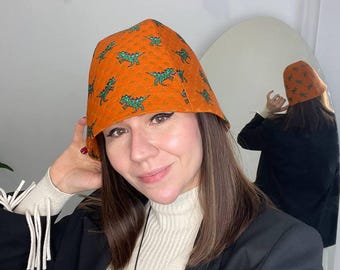 May include: A woman wearing a black blazer and a white turtleneck sweater. She is wearing an orange bucket hat with a green dinosaur print.