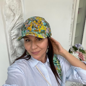 Puede incluir: Gorra de béisbol con estampado floral y bufanda a juego. La gorra presenta un patrón de flores amarillas, azules y verdes. La bufanda está drapeada sobre el hombro. La persona lleva una camisa a rayas azul claro.