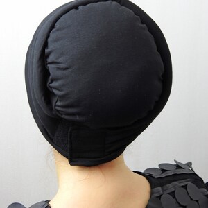 May include: A black head covering with a rounded top and a band that secures with a hook and loop fastener. The head covering is made of a soft, matte fabric. The person is wearing a black top with circular embellishments.