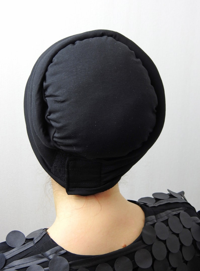 May include: A black head covering with a rounded top and a band that secures with a hook and loop fastener. The head covering is made of a soft, matte fabric. The person is wearing a black top with circular embellishments.