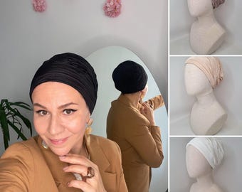 Puede incluir: Una mujer con un turbante negro de estilo turbante y una chaqueta marrón. La mujer mira a la cámara y sonríe. La mujer está de pie frente a un espejo y su reflejo es visible en el espejo. El reflejo de la mujer lleva un turbante negro de estilo turbante y una chaqueta marrón. El reflejo de la mujer mira al espejo y se ajusta el turbante.