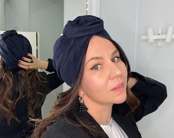 May include: A woman wearing a navy blue turban with a rose-shaped knot on top. She is wearing a black blazer, a white t-shirt with the text "I'm A" on it, and black beaded earrings.