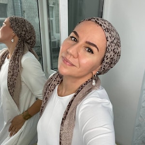 May include: A woman with brown hair is wearing a white shirt and a brown and black patterned headscarf. She is wearing pearl earrings and has a neutral expression on her face.