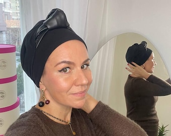 May include: A black turban with a leather bow detail. The turban is worn by a person, paired with a brown sweater and a necklace with cherry-shaped earrings. A mirror reflects the turban and outfit.