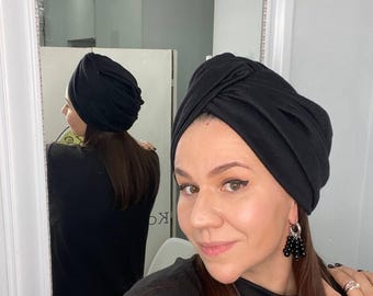 May include: A black turban-style head covering. The head wrap is made of a soft fabric and is worn by a person with long brown hair. The person is wearing a black top and earrings. The image is taken in front of a mirror.