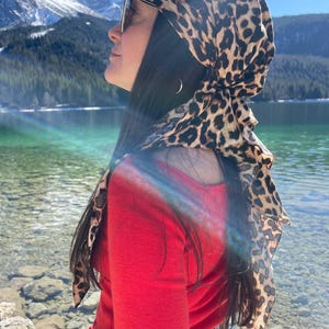 Può includere: Una donna che indossa un maglione rosso e jeans blu con un foulard a stampa leopardata legato intorno alla testa. È in piedi di fronte a un lago con montagne sullo sfondo.