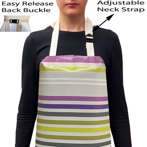 May include: A striped apron with a grey background and purple, white, and green stripes. The apron has a white olive branch design and an adjustable neck strap with a black buckle. The apron has an easy release back buckle.