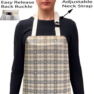 May include: A beige and grey plaid apron with an adjustable neck strap and easy-release back buckle. The apron features a pattern of squares with small heart shapes. The apron is worn over a black long-sleeved shirt and blue jeans.