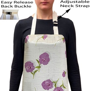 May include: A white apron with a floral pattern featuring purple pansies. The apron has an adjustable neck strap and an easy release back buckle.
