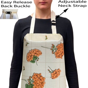 May include: Apron with a floral design featuring orange flowers and green leaves on a light beige background. The apron has an adjustable neck strap and an easy-release back buckle. The apron is worn by a person wearing a black long-sleeve shirt and blue jeans.