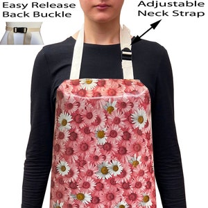 May include: Apron with a pink daisy floral pattern. Features an adjustable neck strap and an easy-release back buckle. The apron is made of a durable material and is designed for kitchen or craft use.