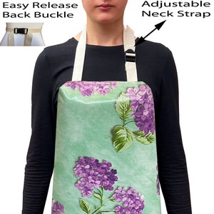 May include: A green apron with a floral print of purple flowers. The apron has an adjustable neck strap and an easy release back buckle.