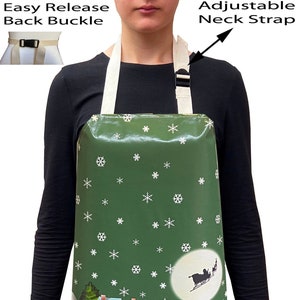 May include: A green Christmas-themed apron with a snowy winter scene, featuring snowmen, houses, and a sleigh pulled by reindeer. The apron has an adjustable neck strap and an easy release back buckle.