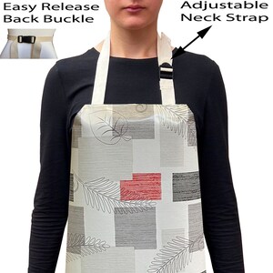 May include: A white apron with a geometric pattern of grey and red squares and black leaf designs. The apron has adjustable neck straps and an easy release back buckle.