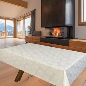 May include: An off-white rectangular tablecloth featuring a delicate floral design. The cloth covers a wooden table with dark brown legs. The scene is a modern living room, complete with a fireplace and large windows overlooking a scenic view.